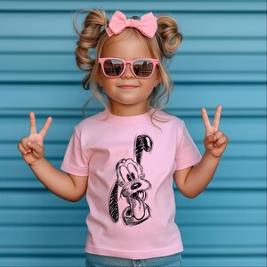 May include: A young child wearing a pink t-shirt featuring a black and white sketch of a cartoon dog. The child is also wearing pink sunglasses and a pink bow. The background is a blue wall.