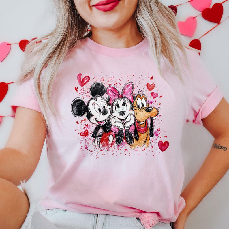 May include: A light pink t-shirt featuring a watercolor design of Mickey Mouse, Minnie Mouse, and Pluto with hearts. The graphic includes red and pink paint splatters, and the characters are smiling. The shirt has short sleeves.