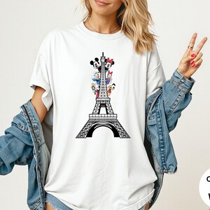 May include: White t-shirt featuring a black Eiffel Tower design with cartoon characters. The design includes Mickey Mouse, Minnie Mouse, Donald Duck, Daisy Duck, and Goofy. The shirt is labeled "Comfort Colors White".