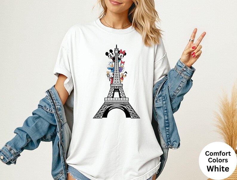 May include: White t-shirt featuring a black Eiffel Tower design with cartoon characters. The design includes Mickey Mouse, Minnie Mouse, Donald Duck, Daisy Duck, and Goofy. The shirt is labeled "Comfort Colors White".