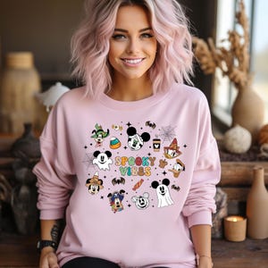 May include: A pink sweatshirt featuring a graphic print of various Disney characters dressed in Halloween costumes. The text "Spooky Vibes" is printed in the centre of the design.