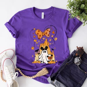 May include: Purple t-shirt with a graphic of a ghost holding a jack-o-lantern in front of a castle with a bow on top. The castle is outlined in black and the ghost is white.
