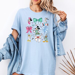 Pu&ograve; includere: T-shirt azzurra con i personaggi dei cartoni animati Topolino e Minnie, Pippo, Paperino e Paperina, insieme a fiocchi e simboli medici. Il design include fiocchi rosa, verdi e bianchi e icone mediche. La maglietta &egrave; in stile casual.