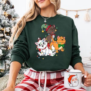 May include: Dark green sweatshirt featuring three cartoon cats decorated with Christmas lights. One cat is white with a red bow, another is brown with an elf hat, and the third is orange with a green bow tie. The person is holding a mug with a Santa Claus design.