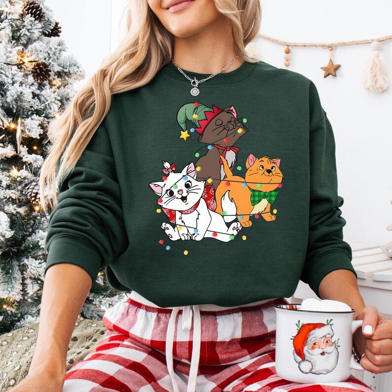 May include: Dark green sweatshirt featuring three cartoon cats decorated with Christmas lights. One cat is white with a red bow, another is brown with an elf hat, and the third is orange with a green bow tie. The person is holding a mug with a Santa Claus design.