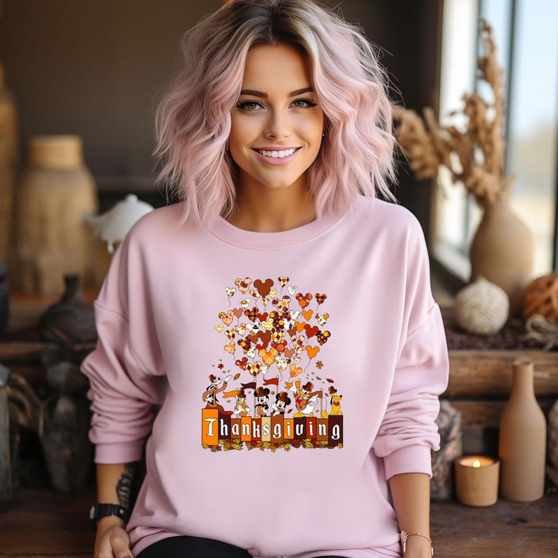 May include: A light pink sweatshirt featuring a Thanksgiving-themed graphic. The graphic includes heart-shaped balloons in shades of brown, orange, and yellow, with the word "Thanksgiving" printed in a rectangular box at the bottom. The sweatshirt has long sleeves.