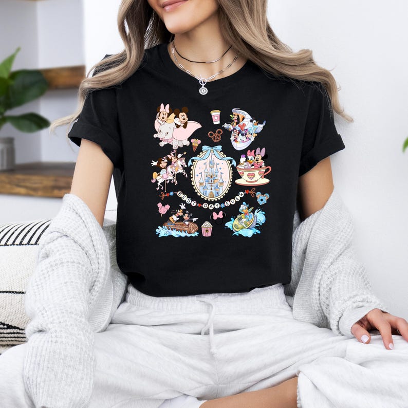 May include: Black t-shirt featuring a colorful graphic of various Disney characters and icons. The design includes images of Minnie and Mickey Mouse, a castle, and other themed elements. The shirt is a classic crew neck style.