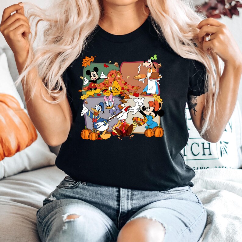May include: Black t-shirt featuring a colorful graphic of Disney characters celebrating Thanksgiving. Mickey Mouse, Donald Duck, Goofy, Minnie Mouse, and other characters are depicted with pumpkins and autumn leaves. The shirt is made of a soft material.