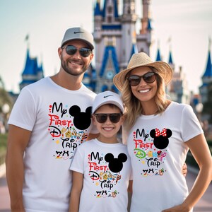 Peut inclure: Trois personnes portant des t-shirts blancs avec un motif Mickey Mouse et le texte "My First Disney Trip 2023". Les chemises ont des graphiques color&eacute;s et une silhouette noire de Mickey Mouse. La famille sourit devant un ch&acirc;teau.