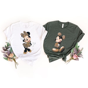 Op de afbeelding: Twee T-shirts met een cartoon afbeelding van Mickey Mouse en Minnie Mouse die luipaardprint outfits dragen. Het Mickey Mouse shirt is groen en het Minnie Mouse shirt is wit.