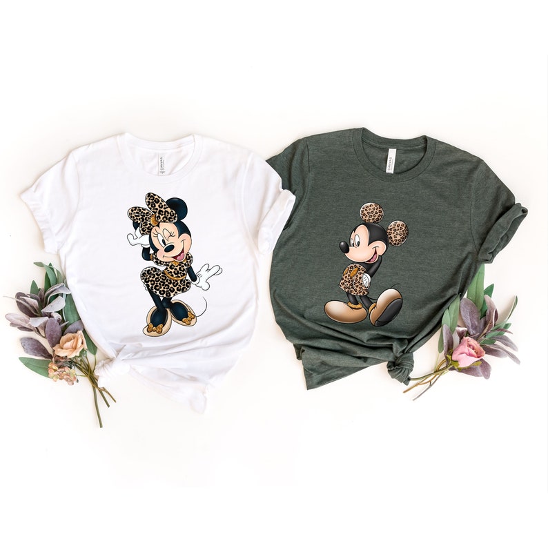 Op de afbeelding: Twee T-shirts met een cartoon afbeelding van Mickey Mouse en Minnie Mouse die luipaardprint outfits dragen. Het Mickey Mouse shirt is groen en het Minnie Mouse shirt is wit.