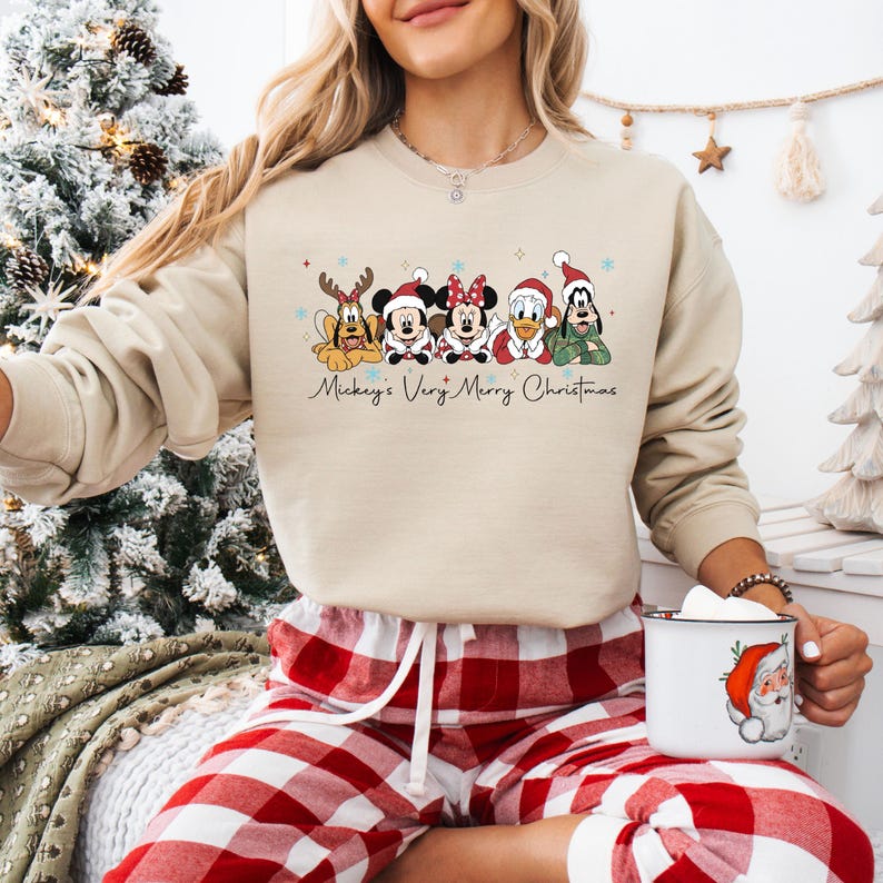 Puede incluir: Sudadera beige con un dise&ntilde;o navide&ntilde;o de personajes de Disney. El texto "Mickey's Very Merry Christmas" est&aacute; escrito debajo. La persona lleva un pantal&oacute;n de pijama a cuadros rojos y blancos y sostiene una taza.