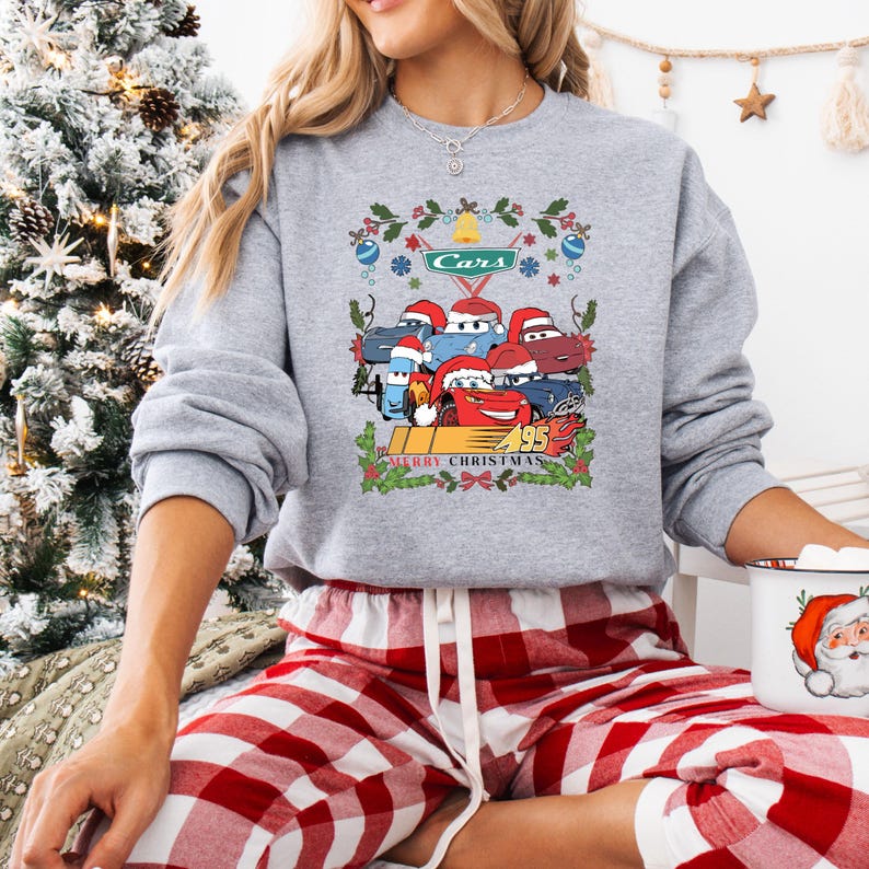 May include: A gray sweatshirt featuring a festive Cars-themed design. The graphic includes cartoon cars wearing Santa hats, the word "Cars", the number "95", and the text "Merry Christmas". The sweatshirt is paired with red and white plaid pajama pants.