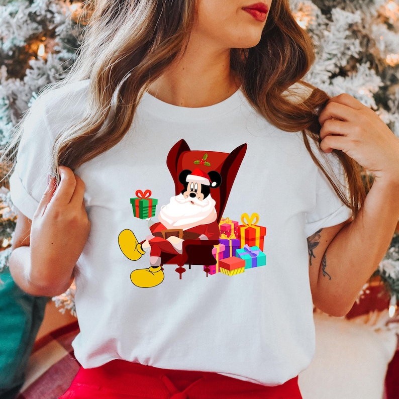 Op de afbeelding: Wit T-shirt met een cartoon van Mickey Mouse als kerstman, zittend in een rode fauteuil. Mickey is omringd door kleurrijk verpakte cadeaus. Het ontwerp bevat een kerstmuts en gele schoenen.