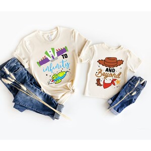 Peut inclure: Deux t-shirts cr&egrave;me avec un th&egrave;me Toy Story. Le plus grand t-shirt pr&eacute;sente une fus&eacute;e de dessin anim&eacute; avec le texte "to infinity" et des &eacute;toiles. Le plus petit t-shirt pr&eacute;sente un chapeau de cow-boy de dessin anim&eacute; avec le texte "and beyond" et une botte de cow-boy.