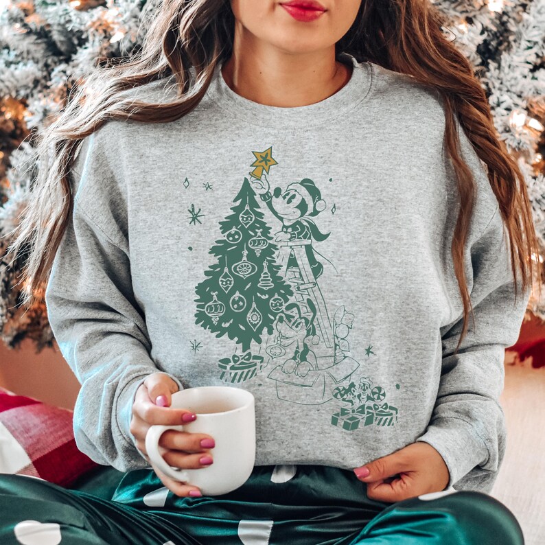 May include: A gray sweatshirt with a vintage-style illustration of Mickey Mouse decorating a Christmas tree. Mickey is on a ladder and holding a star. The illustration is in black and white with green accents.