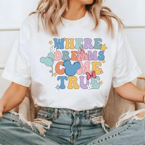May include: White t-shirt with a colorful graphic that says "Where Dreams Come True" with a Mickey Mouse head, a castle, stars, and balloons.
