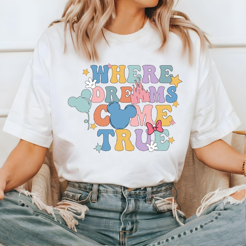 May include: White t-shirt with a colorful graphic that says "Where Dreams Come True" with a Mickey Mouse head, a castle, stars, and balloons.