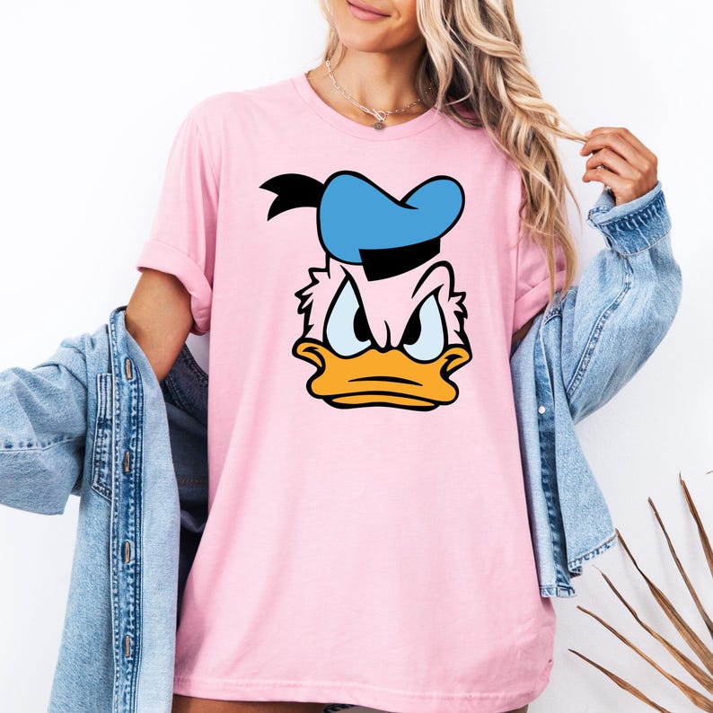 May include: A light pink t-shirt featuring a cartoon image of Donald Duck's face. The design includes a blue sailor hat, an orange beak, and an angry expression. The shirt has short sleeves.