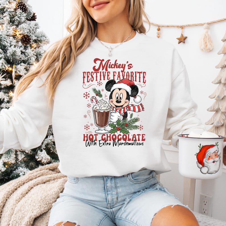 May include: White sweatshirt featuring Mickey Mouse in a Santa hat and scarf, holding a cup of hot chocolate with marshmallows. The text reads "Mickey's Festive Favorite Hot Chocolate With Extra Marshmallows". A Santa mug is also visible.