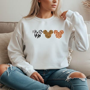May include: White sweatshirt featuring three Mickey Mouse head silhouettes with animal print patterns: zebra, leopard, and giraffe. The sweatshirt has long sleeves and a crew neck. The overall theme is a blend of Disney and safari.