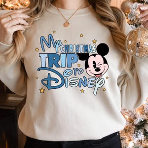Puede incluir: Una sudadera blanca con un dise&ntilde;o gr&aacute;fico azul y amarillo con Mickey Mouse y el texto "My Christmas Trip to Disney".