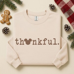 Puede incluir: Sudadera beige de cuello redondo con la palabra "thankful" en letras marrones. La "o" de la palabra se reemplaza con una cabeza de Mickey Mouse. La sudadera est&aacute; rodeada de pi&ntilde;as, vegetaci&oacute;n y un hombre de jengibre.