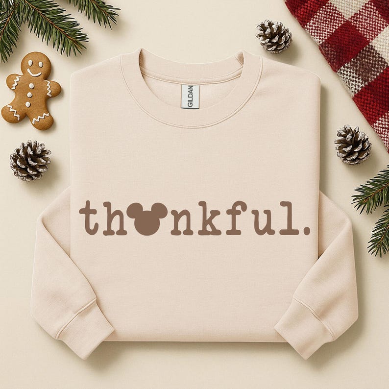 Puede incluir: Sudadera beige de cuello redondo con la palabra "thankful" en letras marrones. La "o" de la palabra se reemplaza con una cabeza de Mickey Mouse. La sudadera est&aacute; rodeada de pi&ntilde;as, vegetaci&oacute;n y un hombre de jengibre.