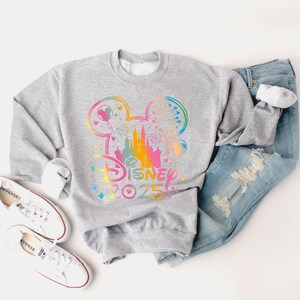 May include: A gray sweatshirt with a rainbow-colored graphic of Mickey Mouse ears, a castle, and fireworks. The text "Disney 2025" is printed below the graphic.