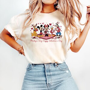 May include: Cream-colored t-shirt featuring a graphic of Disney characters, including Mickey and Minnie Mouse, celebrating Valentine's Day. The design includes hearts and the text "Mickey's Very Happy Valentine's Day" in a pink script.