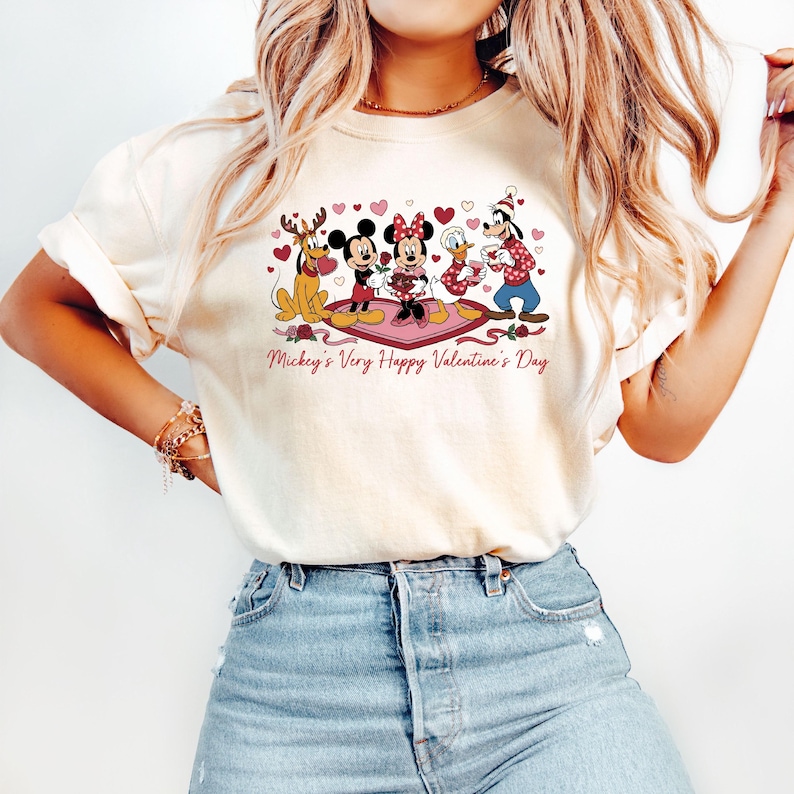 May include: Cream-colored t-shirt featuring a graphic of Disney characters, including Mickey and Minnie Mouse, celebrating Valentine's Day. The design includes hearts and the text "Mickey's Very Happy Valentine's Day" in a pink script.