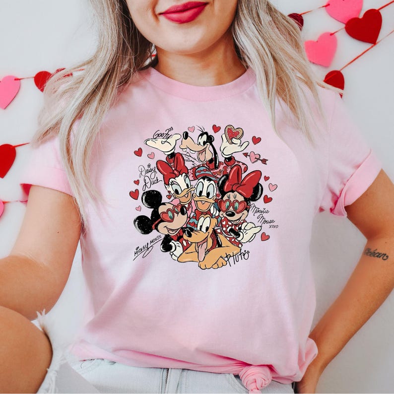 May include: A light pink t-shirt featuring a graphic of Disney characters including Mickey Mouse, Minnie Mouse, Donald Duck, Daisy Duck, Goofy, and Pluto, surrounded by hearts. The shirt has the text "Mickey Mouse" and "Minnie Mouse" in cursive.