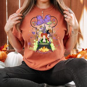 May include: A burnt orange t-shirt with a graphic of the Sanderson sisters from the movie Hocus Pocus. The graphic features the sisters standing in front of a cauldron with a castle in the background. The graphic is surrounded by hearts, stars, and fireworks. The text "Hocus Pocus" is written below the graphic.
