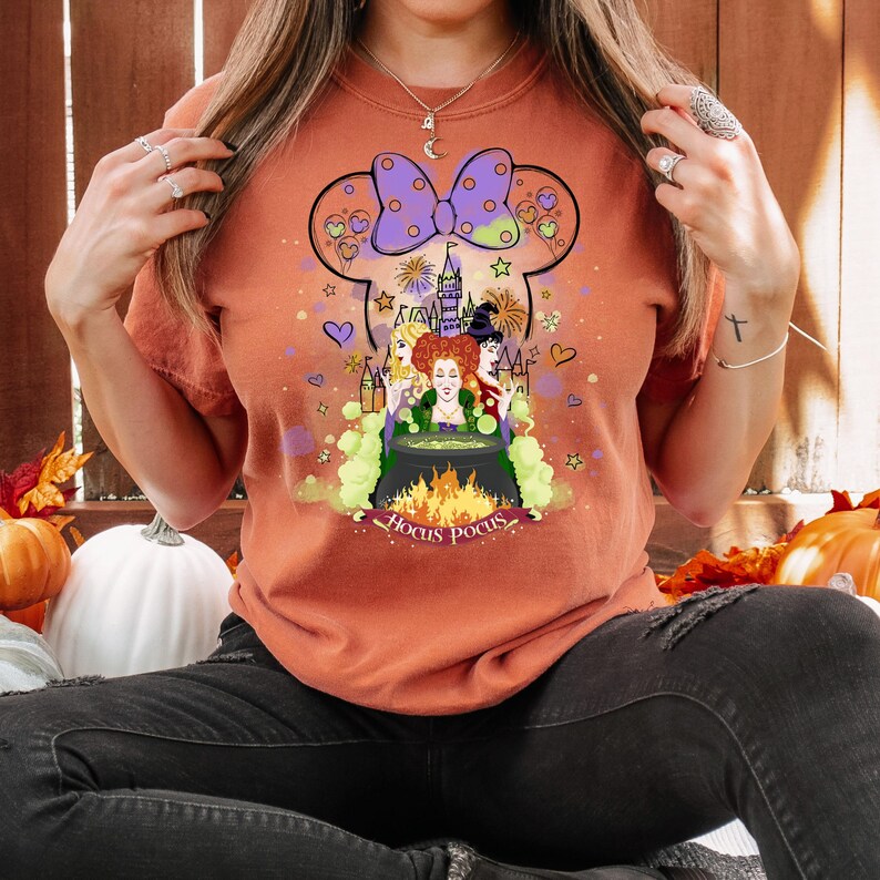 May include: A burnt orange t-shirt with a graphic of the Sanderson sisters from the movie Hocus Pocus. The graphic features the sisters standing in front of a cauldron with a castle in the background. The graphic is surrounded by hearts, stars, and fireworks. The text "Hocus Pocus" is written below the graphic.