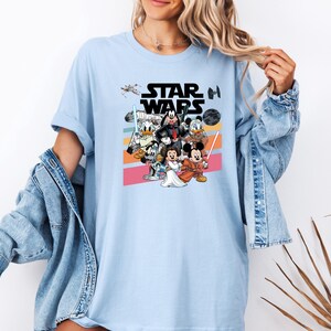 K&ouml;nnte beinhalten: Hellblaues T-Shirt mit einer Grafik von Disney-Figuren in Star Wars-Kleidung. Das Design umfasst Micky Maus, Minnie Maus, Donald Duck und Goofy, mit dem Star Wars-Logo dar&uuml;ber. Das Shirt hat einen Rundhalsausschnitt und kurze &Auml;rmel.