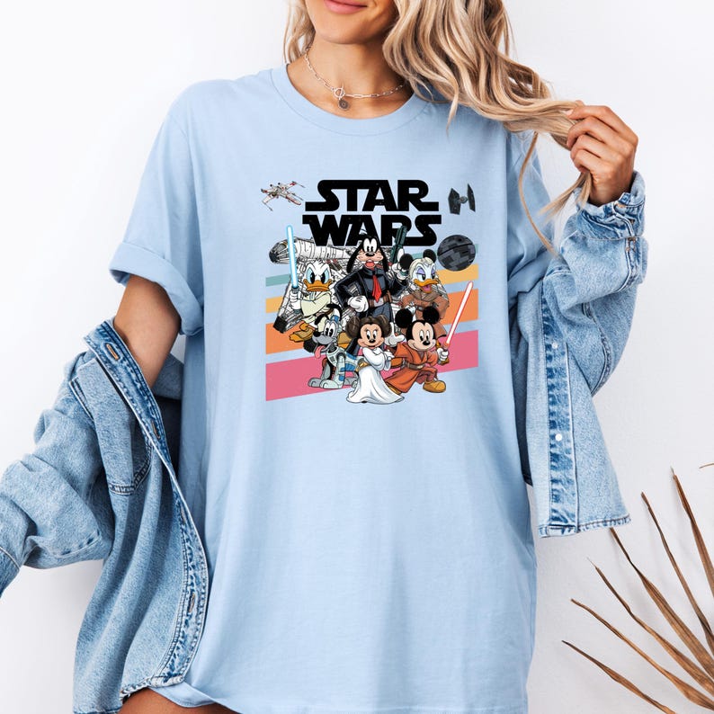 K&ouml;nnte beinhalten: Hellblaues T-Shirt mit einer Grafik von Disney-Figuren in Star Wars-Kleidung. Das Design umfasst Micky Maus, Minnie Maus, Donald Duck und Goofy, mit dem Star Wars-Logo dar&uuml;ber. Das Shirt hat einen Rundhalsausschnitt und kurze &Auml;rmel.