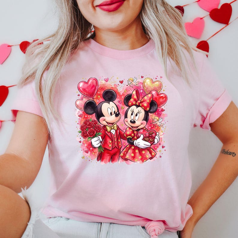 May include: Pink t-shirt featuring a graphic of Mickey and Minnie Mouse in Valentine's Day attire. Mickey holds roses, and Minnie holds a heart-shaped box. Both are surrounded by heart-shaped balloons and a heart-filled background.