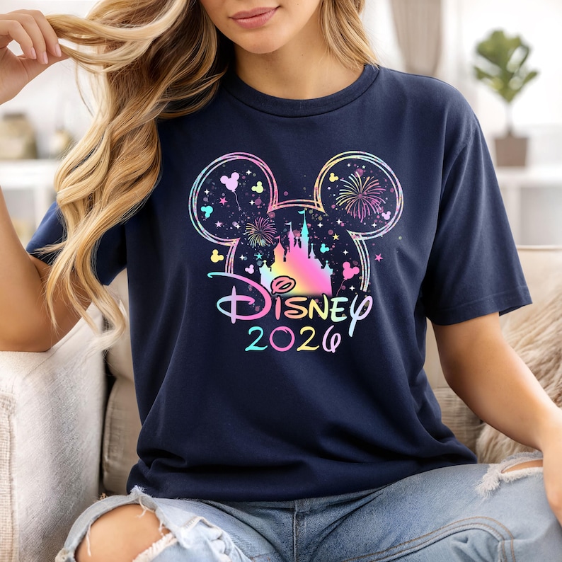 May include: Navy blue t-shirt featuring a colorful Disney design with Mickey Mouse ears, a castle, fireworks, and the text "Disney 2026". The design incorporates pastel colors and a whimsical aesthetic.