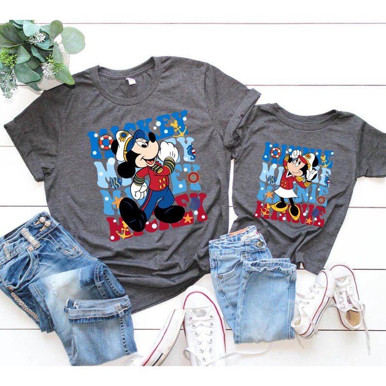 May include: Two grey t-shirts with a nautical theme featuring Mickey Mouse and Minnie Mouse. The shirts have a blue, red, and white color scheme with anchors, stars, and waves. The text on the shirts reads "Mickey Mouse" and "Minnie Mouse".