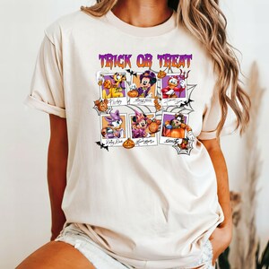 May include: Beige t-shirt featuring a Halloween-themed graphic. The design includes the text "Trick or Treat" in purple above six framed images of Disney characters dressed in Halloween costumes, including Mickey Mouse, Pluto, and Donald Duck.