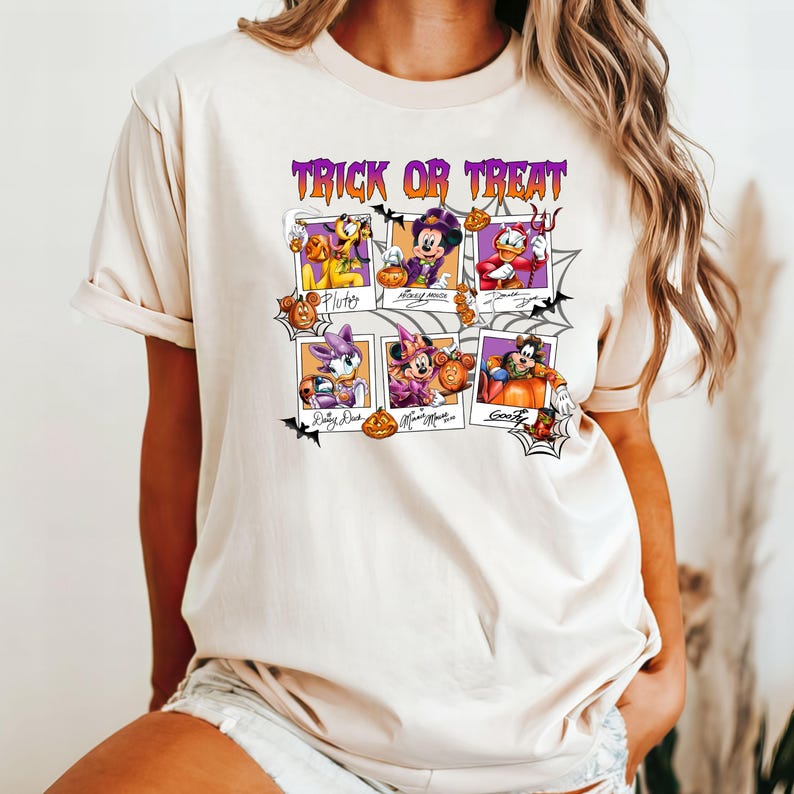 May include: Beige t-shirt featuring a Halloween-themed graphic. The design includes the text "Trick or Treat" in purple above six framed images of Disney characters dressed in Halloween costumes, including Mickey Mouse, Pluto, and Donald Duck.