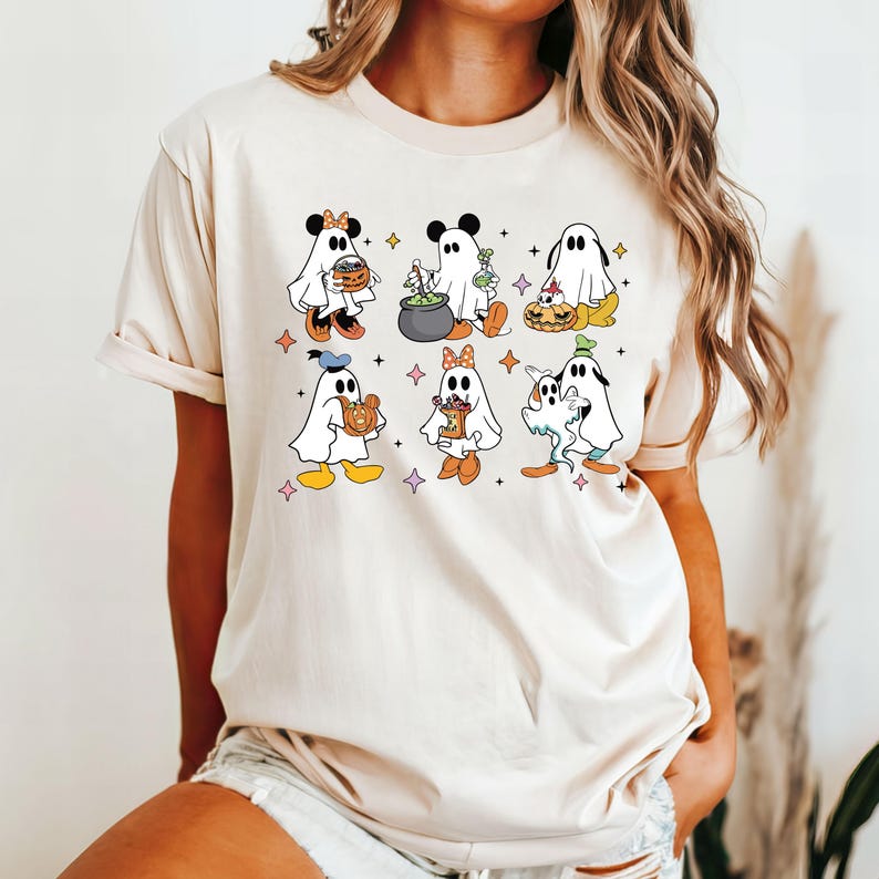 May include: A white t-shirt featuring a graphic of six cartoon ghosts dressed in Halloween costumes. The ghosts are holding various Halloween-themed items, such as a cauldron, a pumpkin, and a candy bucket.