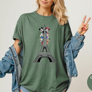 May include: Moss green t-shirt featuring a black and white Eiffel Tower design with cartoon characters. The shirt has short sleeves and a relaxed fit. The words "Comfort Colors Moss" are printed on a green circle.