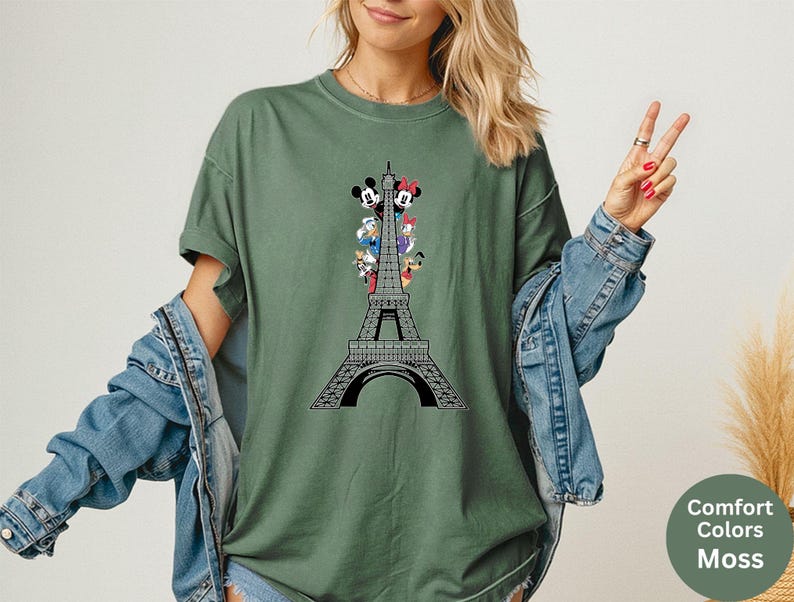 May include: Moss green t-shirt featuring a black and white Eiffel Tower design with cartoon characters. The shirt has short sleeves and a relaxed fit. The words "Comfort Colors Moss" are printed on a green circle.