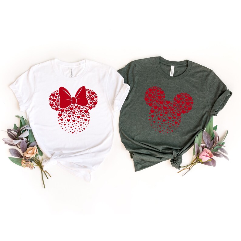 May include: Two t-shirts with a red heart Mickey Mouse design. The shirt on the left is white and the shirt on the right is dark green.