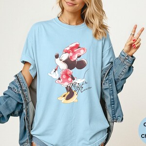 K&ouml;nnte beinhalten: Hellblaues T-Shirt mit Minnie Mouse-Grafik. Die Zeichentrickfigur tr&auml;gt ein rotes Kleid mit Tupfen und eine Schleife, mit dem Schriftzug "Minnie Mouse" in Schreibschrift. Teil der Comfort Colors Chambray-Kollektion.