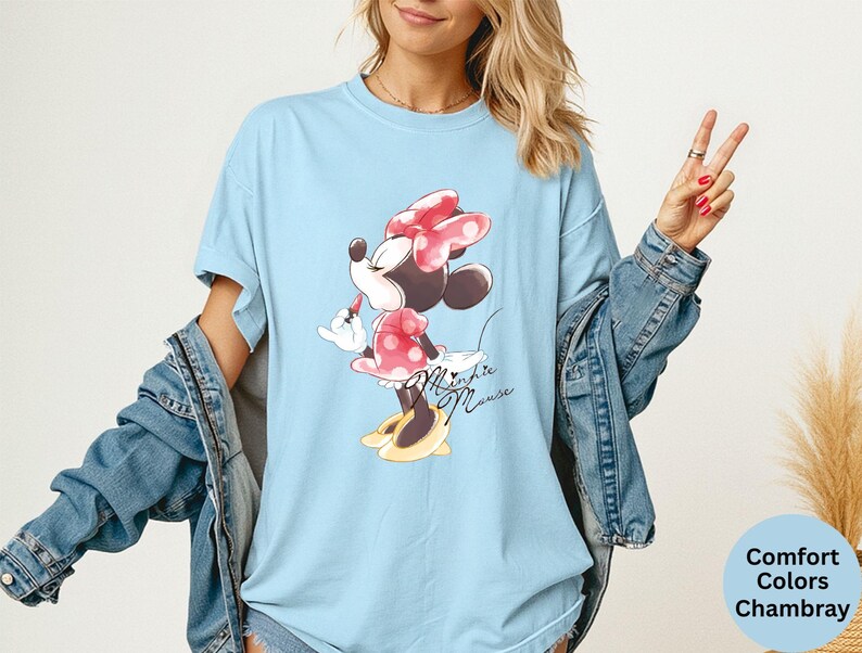 K&ouml;nnte beinhalten: Hellblaues T-Shirt mit Minnie Mouse-Grafik. Die Zeichentrickfigur tr&auml;gt ein rotes Kleid mit Tupfen und eine Schleife, mit dem Schriftzug "Minnie Mouse" in Schreibschrift. Teil der Comfort Colors Chambray-Kollektion.