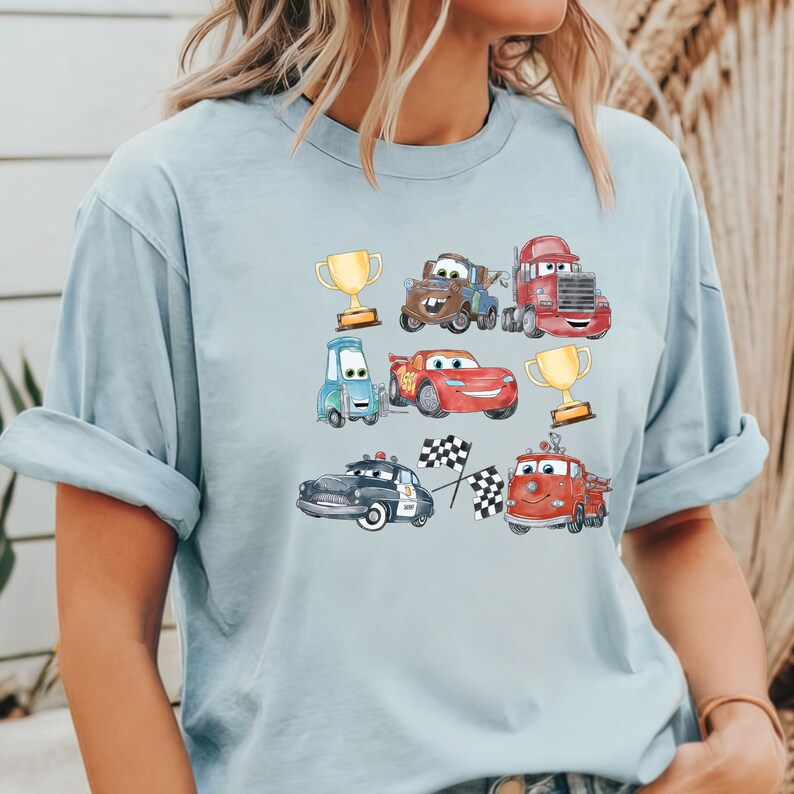 May include: A light blue t-shirt with a cartoon design featuring various cars from the Cars movie franchise. The cars are depicted in a watercolor style and include Lightning McQueen, Mater, and other characters. The design also includes checkered flags and trophies.