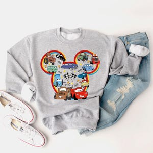 May include: Gray sweatshirt with a Mickey Mouse head design featuring various Cars characters, including Mater, Lightning McQueen, and other vehicles. The design is in a colorful cartoon style.