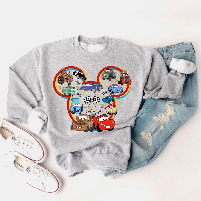May include: Gray sweatshirt with a Mickey Mouse head design featuring various Cars characters, including Mater, Lightning McQueen, and other vehicles. The design is in a colorful cartoon style.
